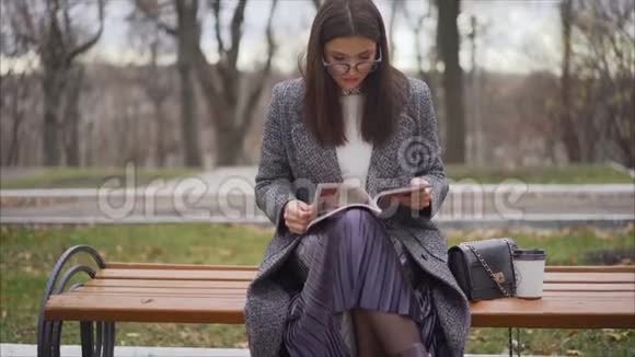 秋天的城市里年轻漂亮的商业女人坐在长凳上秋天美丽的女孩坐在长凳上树叶上视频的预览图