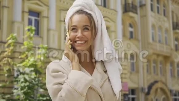一幅美丽的微笑女孩穿着浴袍头上有毛巾在街上用手机说话自信的女孩视频的预览图