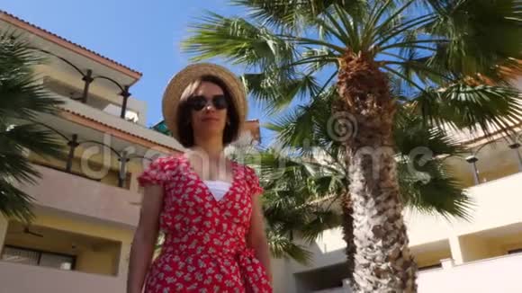 年轻迷人的女人享受阳光戴着帽子和太阳镜背景上有棕榈树酒店花园斯隆视频的预览图