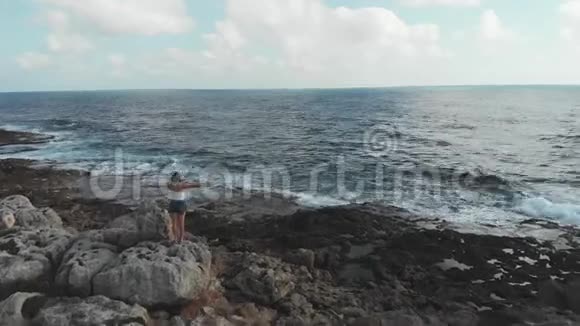 女人的轮廓与手广泛的空中岩石悬崖沿着海洋和波浪冲击海岸线张开双臂的女性视频的预览图