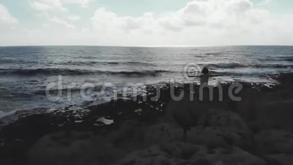 黑色的女性轮廓手臂张开站在悬崖上强烈的波浪撞击海洋海滩空中无人机视频的预览图
