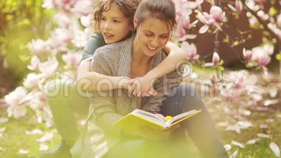 幸福的家庭在春天的草地上以粉红色的玉兰为背景母亲和儿子坐在上面读一本书视频的预览图