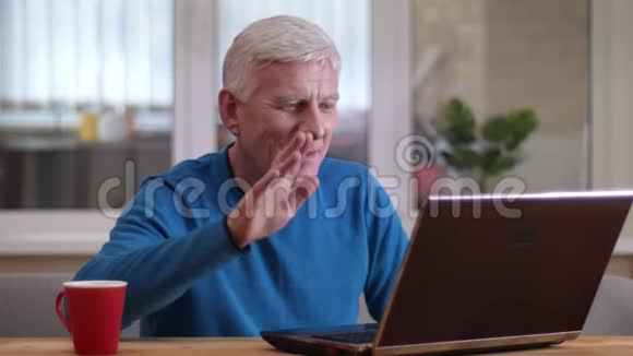 在一个舒适的公寓里一个老白种人在笔记本电脑上打着视频电话在室内开心地笑着的特写镜头视频的预览图