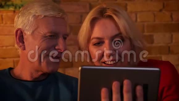 在平板电脑上播放视频的一对幸福的老夫妇的特写镜头笑着坐在室内的沙发上视频的预览图