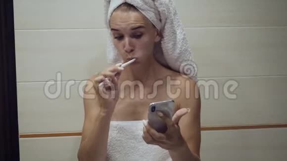年轻的女人刷牙看着智能手机视频的预览图