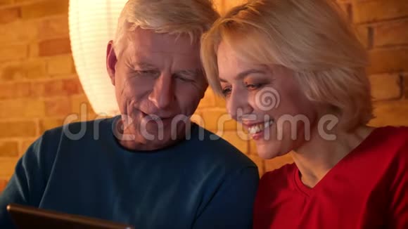 在平板电脑上看喜剧的一对老年幸福夫妇的特写镜头欢快地坐在室内的沙发上视频的预览图