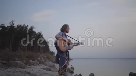 在夏日海滩附近的傍晚五颜六色的日落背景下嬉皮士年轻人在弹声吉他视频的预览图