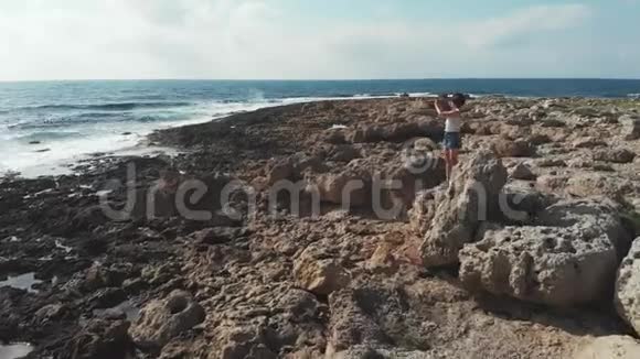岩石上的年轻女子用照相机拍照强烈的海浪冲击海岸线空中无人机视野塞浦路斯日落视频的预览图
