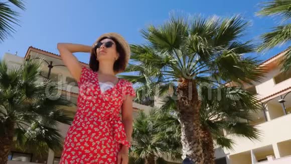 年轻迷人的可爱摆姿势穿着红色连衣裙和太阳镜背景上有蓝天和棕榈树慢动作视频的预览图