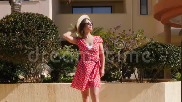 美丽的白种人女人戴着太阳镜戴着帽子穿着红色的连衣裙带着棕榈树走向花园里的相机慢一点视频的预览图