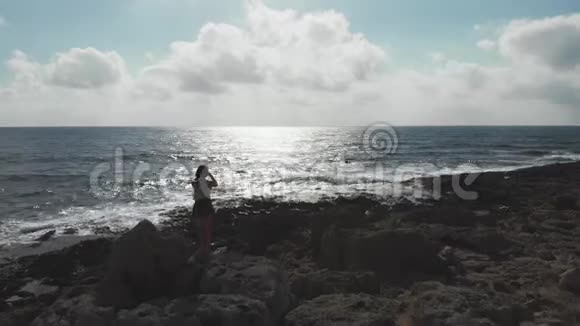空中无人机拍摄妇女与电话相机站在岩石海滩拍摄太阳路和地平线海洋海岸带视频的预览图