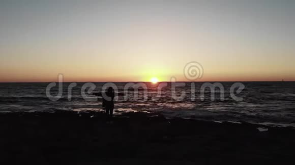 落基海滩上女人的黑色剪影在日落时在海洋上蔓延海浪撞击海滩空中无人驾驶飞机视频的预览图