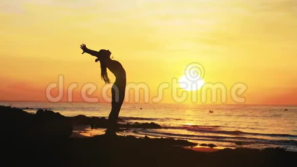 年轻女子的剪影正在日落时在海边做瑜伽练习视频的预览图