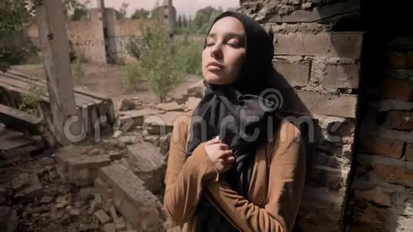 年轻的穆斯林妇女戴着头巾站在被毁的砖楼旁看着摄像机悲伤和沮丧视频的预览图
