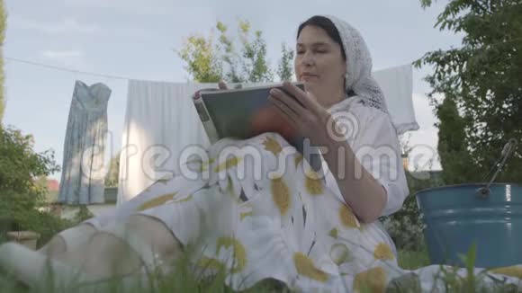 漂亮的高级妇女头上戴着一条白色披肩坐在花园前的草地上休息视频的预览图