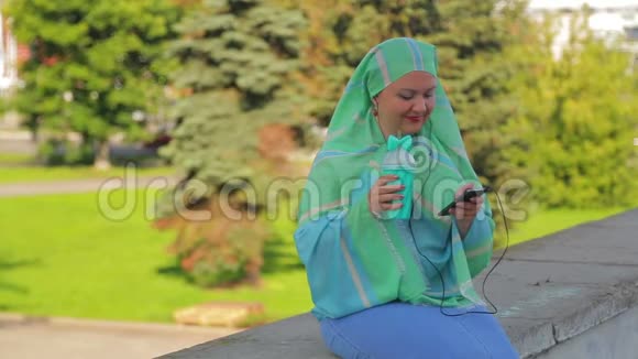 公园里一个戴着浅色围巾的年轻活泼的穆斯林妇女正在喝咖啡戴着耳机听信者说话视频的预览图