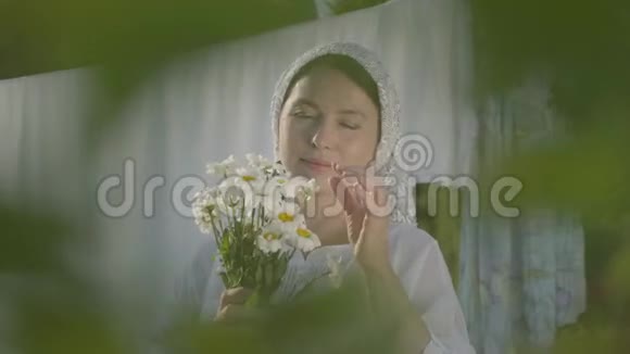 漂亮的老年妇女头上戴着白色披肩在户外晾衣绳上撕下雏菊花瓣洗衣日视频的预览图