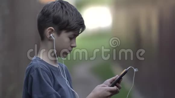 戴着耳机的男孩站在一个带电话的围栏附近环顾四周在户外玩游戏视频的预览图