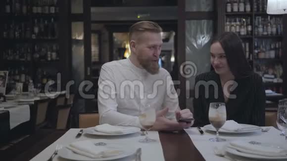 漂亮的胡须男和漂亮的黑发女人坐在餐桌旁的餐馆里那个男人给他女朋友看视频的预览图