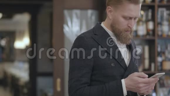 一位成功的年轻有胡子自信的商人站在餐馆或餐馆里用手机打字的肖像视频的预览图