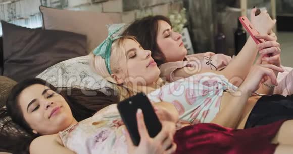 漂亮的多民族女性会穿着睡衣躺在床上用智能手机消费视频的预览图