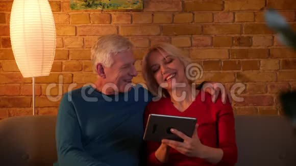 一对老年夫妇在平板电脑上浏览社交媒体的特写镜头他们在室内舒适地坐在沙发上开心地笑着视频的预览图