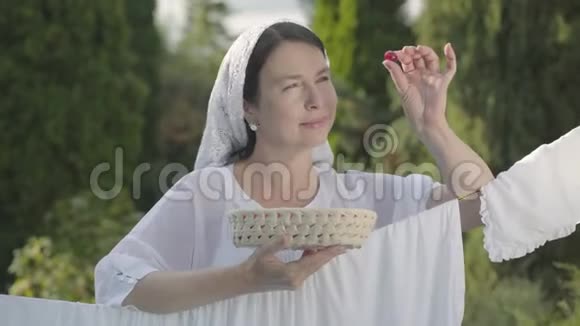 美丽的女人头上戴着白色披肩吃着樱桃看着相机微笑着在户外的晾衣绳上视频的预览图