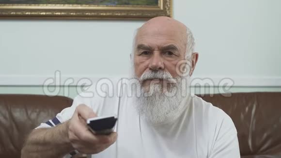 肖像成熟的老人穿着白色t恤坐在棕色皮革沙发上看电视老人打开通道视频的预览图