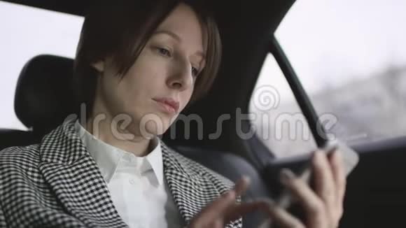 穿着白色上衣的棕色头发的漂亮女人在车上检查外套在手机上输入信息视频的预览图