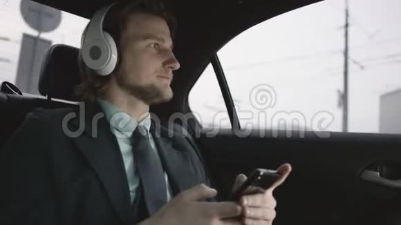 漂亮的男人棕色头发穿着开心果衬衫穿着灰色西装坐在车里通过手机输入信息听音乐视频的预览图