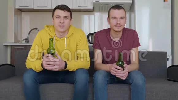 两个人坐在沙发上看电视上的足球赛手里拿着啤酒瓶视频的预览图