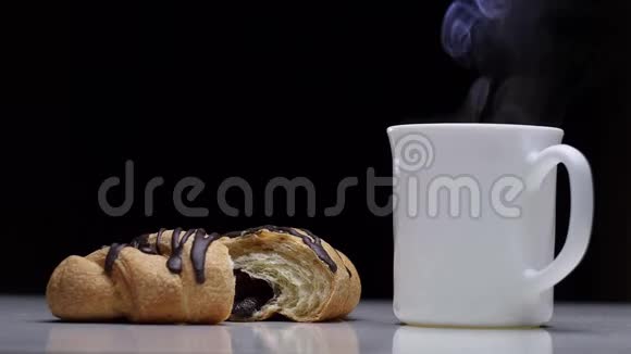 新鲜巧克力牛角面包和一杯带蒸汽的热咖啡放在黑色背景的白色桌子上视频的预览图