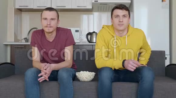 两个男人坐在沙发上用爆米花碗看电视上的足球赛视频的预览图