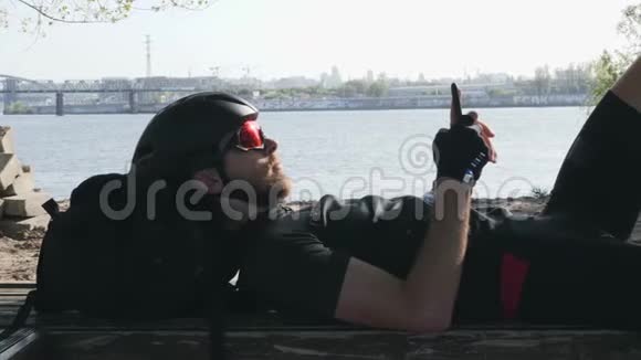 骑自行车的人穿着黑色运动服坐在长凳上手里拿着手机太阳在太阳镜里反射带着头盔的线人用他的视频的预览图