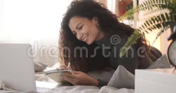 在床上使用笔记本电脑和书写笔记的女人视频的预览图