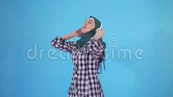 一位带着耳机听音乐和在蓝色背景下跳舞的年轻穆斯林妇女的肖像视频的预览图