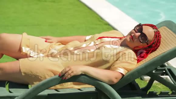 一位年轻女子穿着浴袍在游泳池附近的躺椅上打日光浴视频的预览图