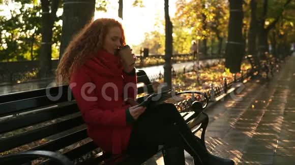 穿红色衣服的女人在秋天的天气里喝咖啡坐在公园的长凳上用平板电脑红头女孩视频的预览图