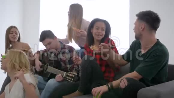 年轻人玩得开心坐在沙发上喝比萨饼快乐的朋友在家庭聚会上用吉他唱歌视频的预览图