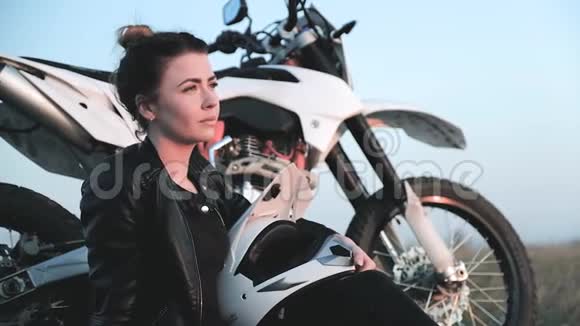 手里拿着摩托车头盔的年轻女孩在摩托车旁享受日落视频的预览图