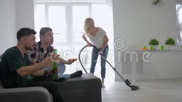 体育迷们拿着啤酒和披萨在沙发上看足球然后坐在女孩的背景上用吸尘器打扫视频的预览图
