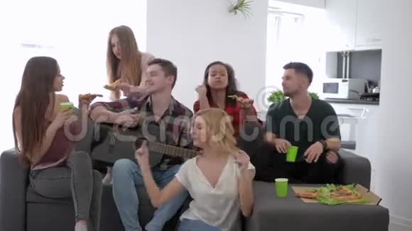 朋友们用吉他唱歌在家里的聚会上坐在沙发上吃披萨友谊和关系视频的预览图