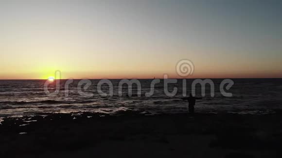 黑黝黝的人影双臂张开站在海滩上望着海面上的日落小船在水平线上视频的预览图