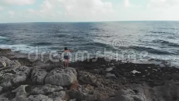 年轻的白种人女人站在巨大的岩石上双臂张开看着暴风雨的海洋海浪冲击着海岸空中飞行视频的预览图