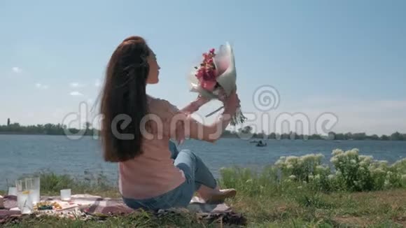 在阳光明媚的天气里在河边的草坪上野餐快乐的长发女子在大自然中享受着美丽的花束视频的预览图