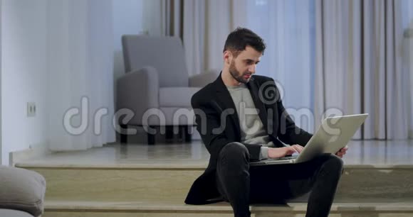 年轻的有魅力的男人穿着黑色西装坐在家里感到快乐和伟大他合上了笔记本视频的预览图