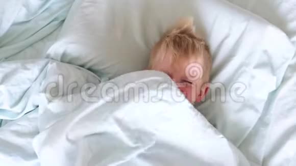 一个金发小男孩躺在床上微笑着玩得很开心躲在被子下面从他下面偷看视频的预览图