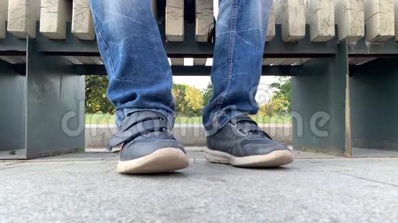 一个无聊的少年坐在城市公园的长凳上穿着牛仔裤和鞋子他的腿很懒过马路视频的预览图