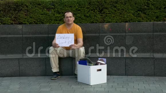 街上的失业男子带着刻字的科顿需要工作被解雇的人坐在一条水泥街上视频的预览图