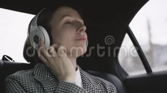 漂亮的女人棕色头发穿着白色衬衫在车上检查外套戴着耳机听音乐视频的预览图